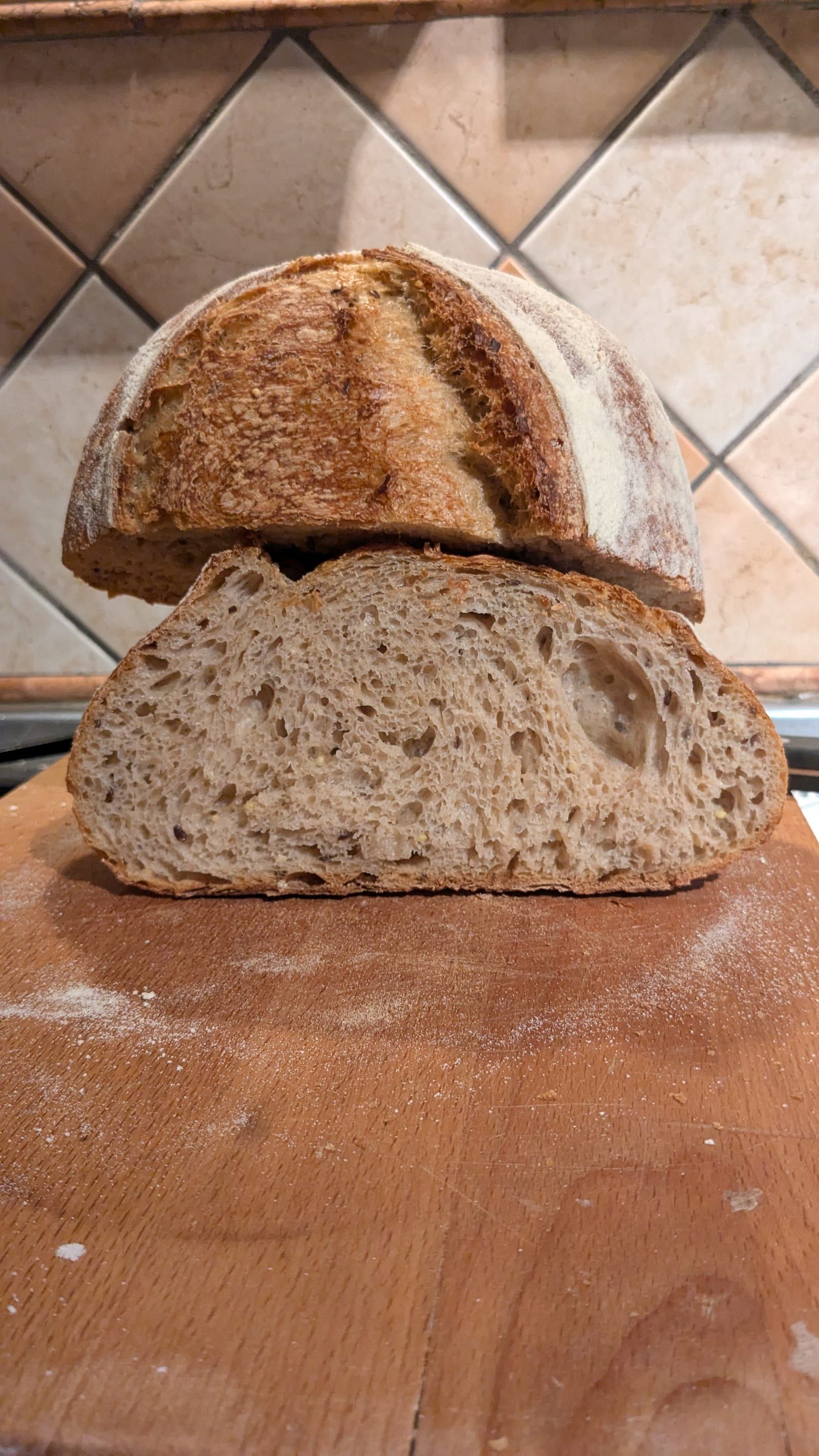 A halved sourdough loaf, crust stacked on crumb, resting on a floured wooden board against a tiled wall.
